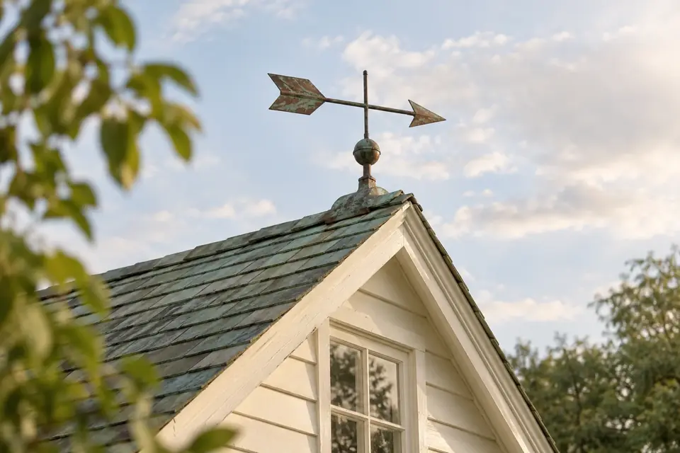 A copper weathervane on the ridge of a cottage roof against a pale sky